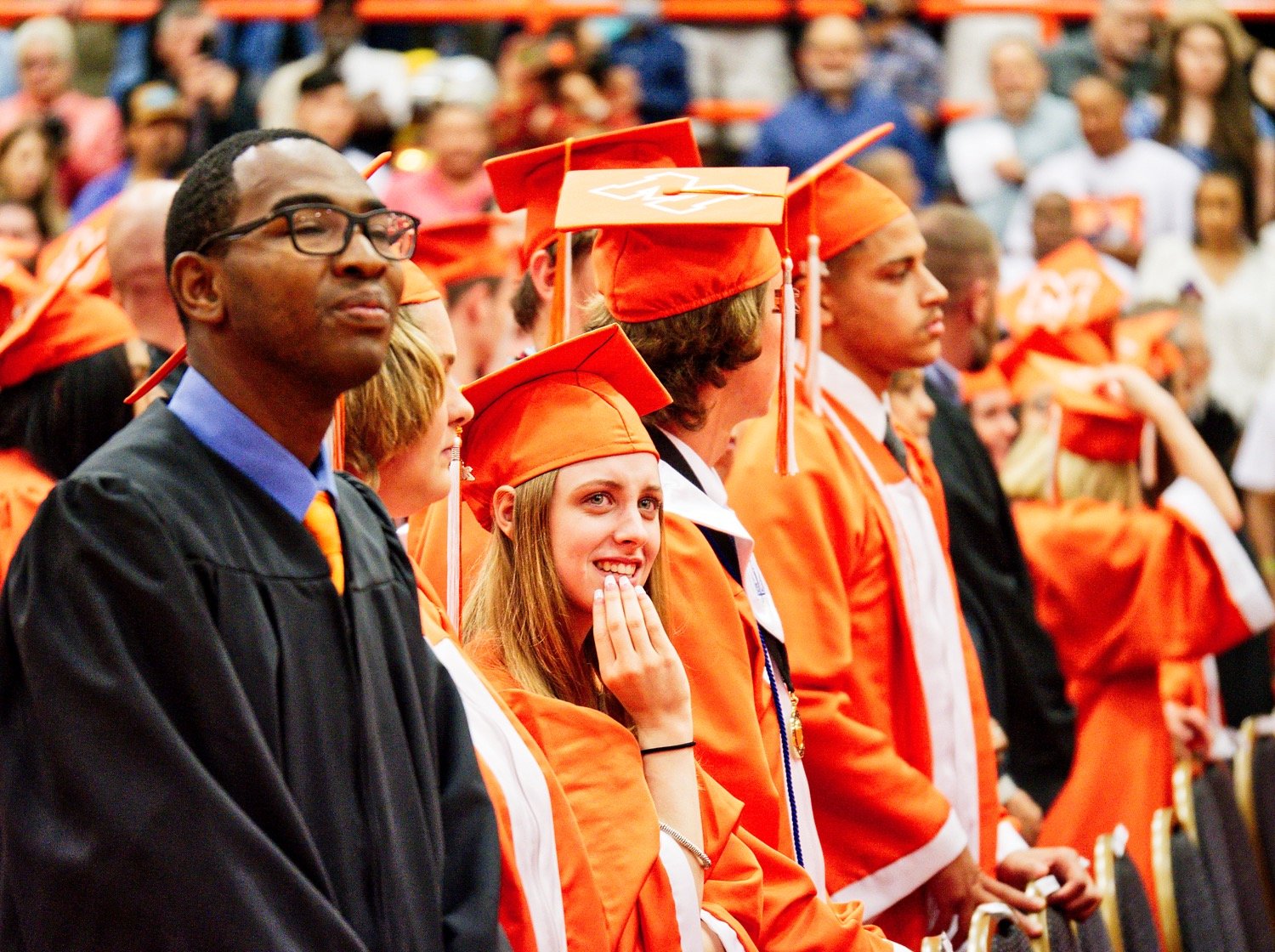 2021 grad Austin Hill spots loved ones in the crowd filling the Mineola High gym just before the commencement ceremonies begin. At left, coach Jaylon Harper, MHS class of 2020, looks on with pride during his first ceremony as staff since graduating. [more moments of Mineola grads]